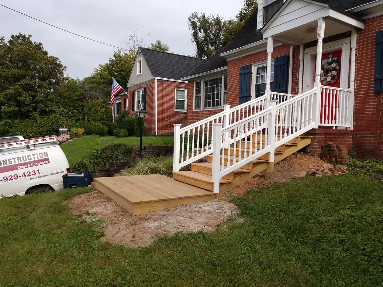 New wood steps and landing with white railings leading to the red front door of a brick house
